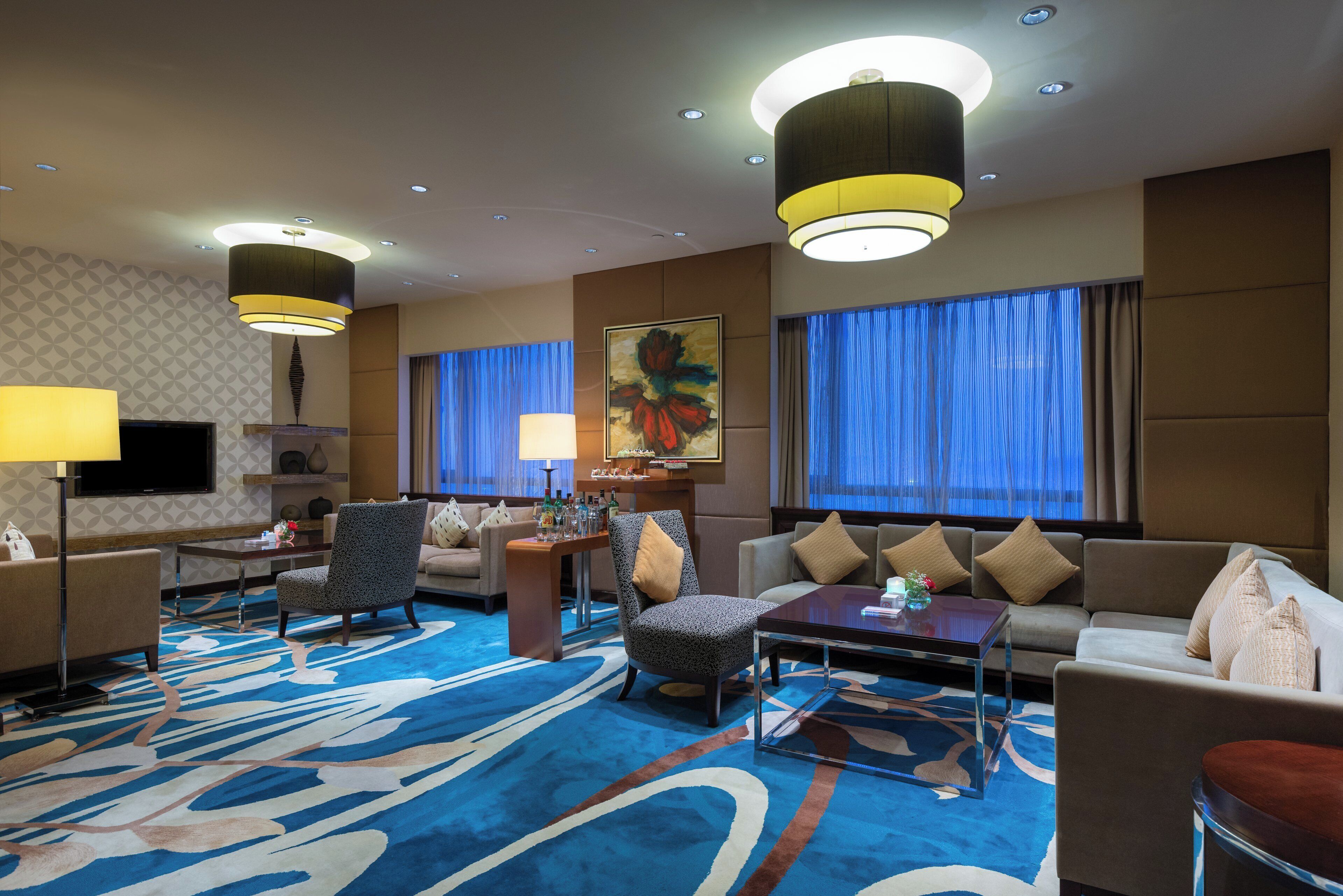 A hotel room with a large blue patterned carpet, a sofa set, a coffee table, a TV, and a painting on the wall.