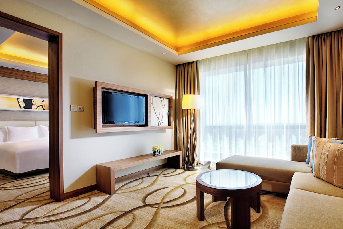 A hotel room with a bed, a sofa, a TV mounted on the wall, a lamp, and a large window with curtains.