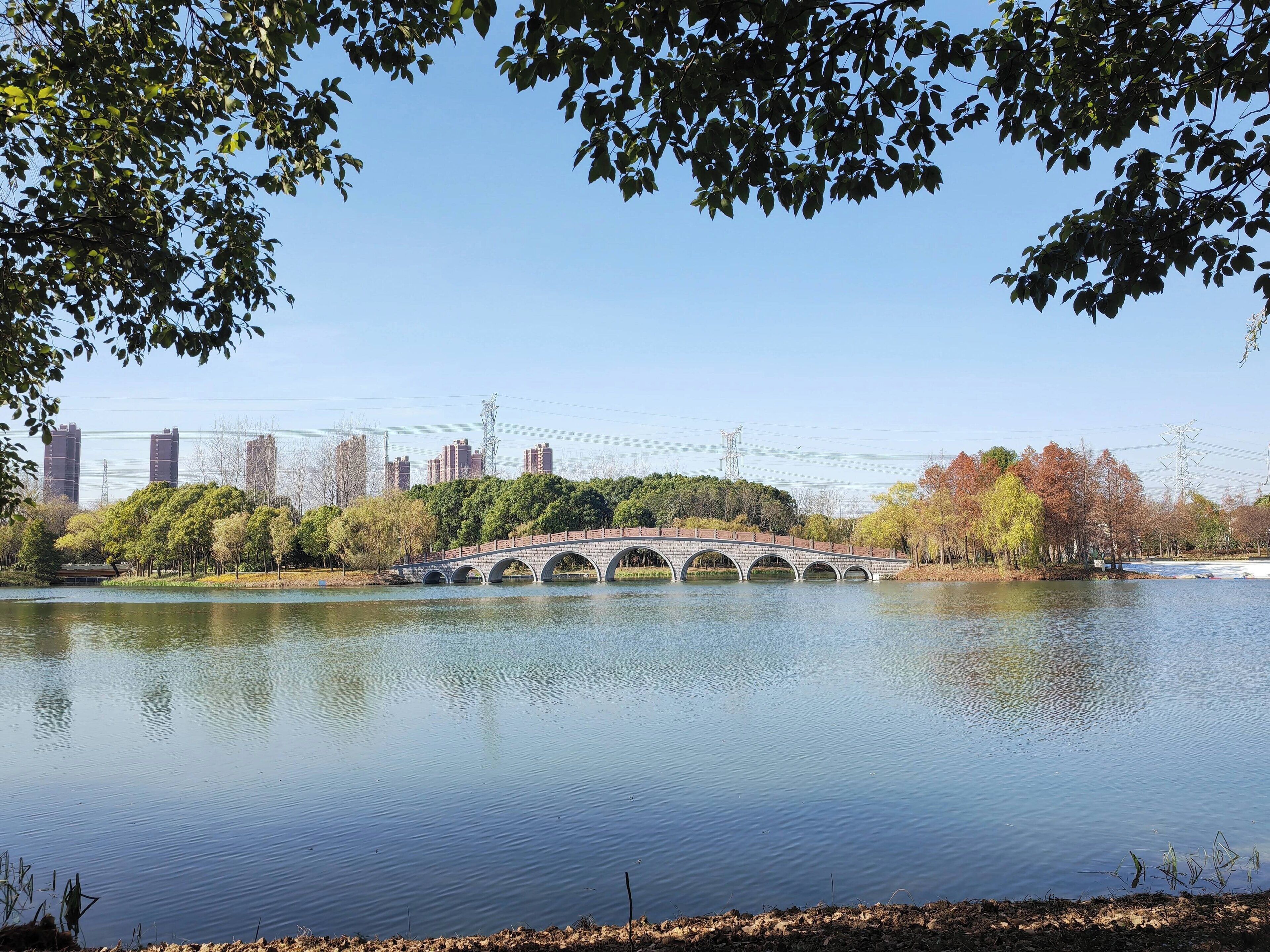 A bridge over a calm lake with trees and buildings in the background.