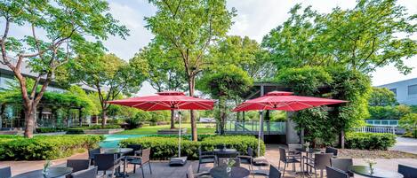 An outdoor patio area with round tables and chairs, red umbrellas, and a pool in the background.