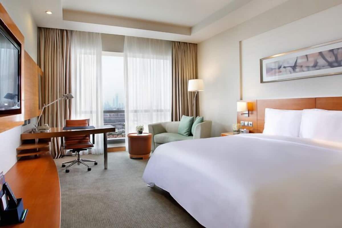 A hotel room with a large bed, a desk, a chair, a lamp, and a view of the city.