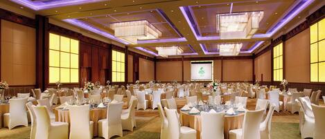 Une grande salle de banquet avec des tables rondes, des chaises blanches et un écran de projection à l’arrière-plan.