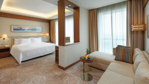 Quarto de hotel com uma cama, um sofá, uma mesinha com frutas, um quadro na parede e uma janela com cortinas.