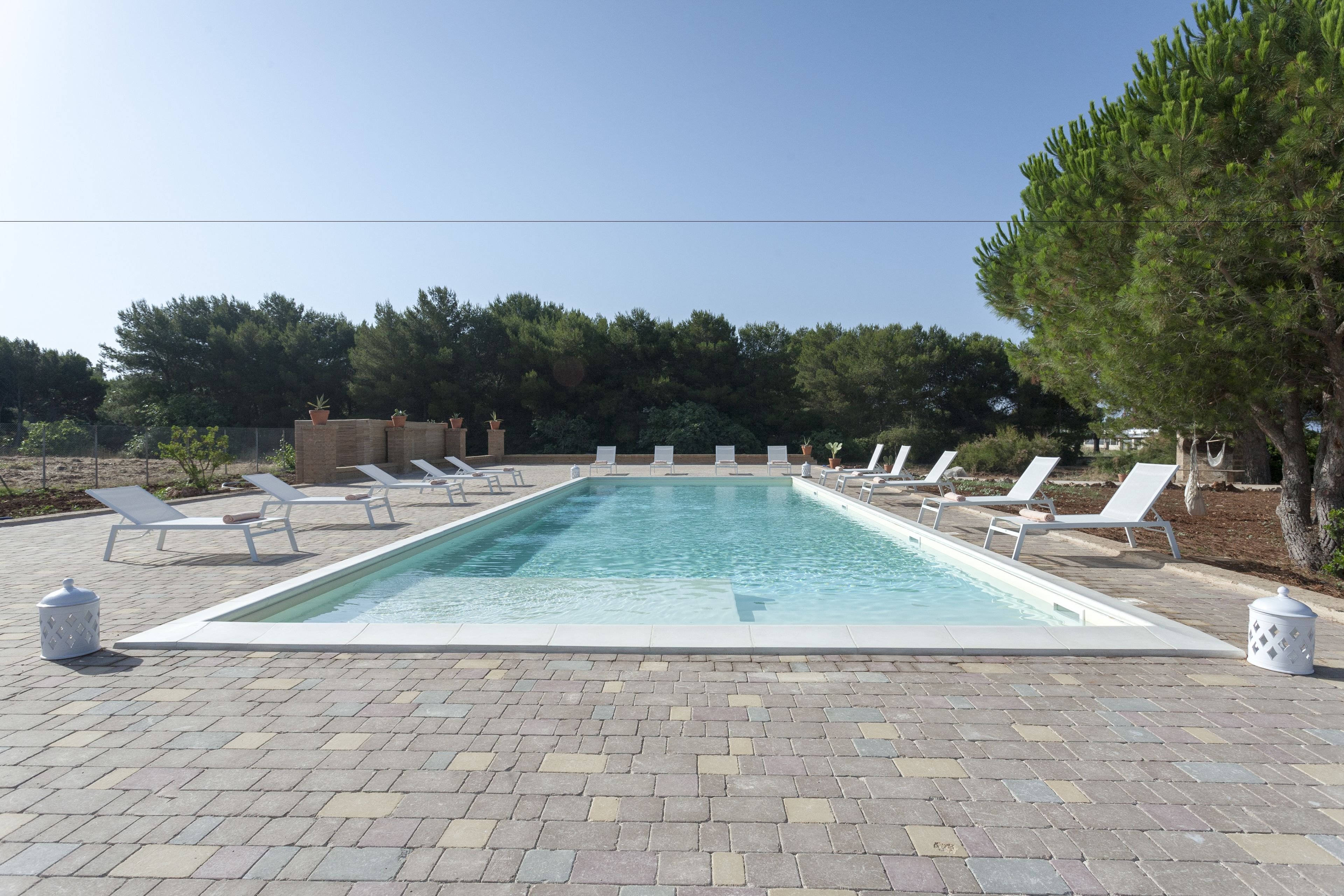 A rectangular swimming pool with white lounge chairs around it, surrounded by a paved area and trees.