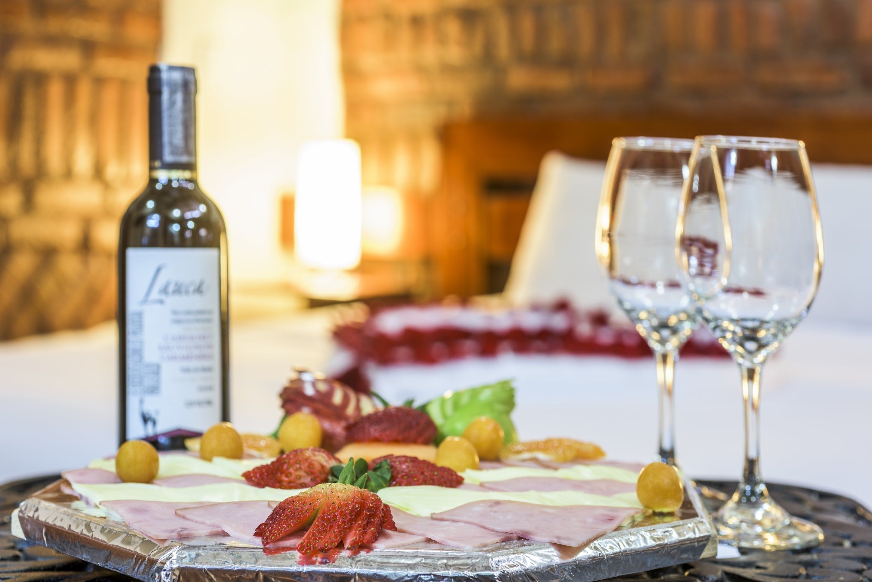 Una tabla de fiambres con diferentes carnes y frutas, dos copas de vino y una botella sobre una mesa.
