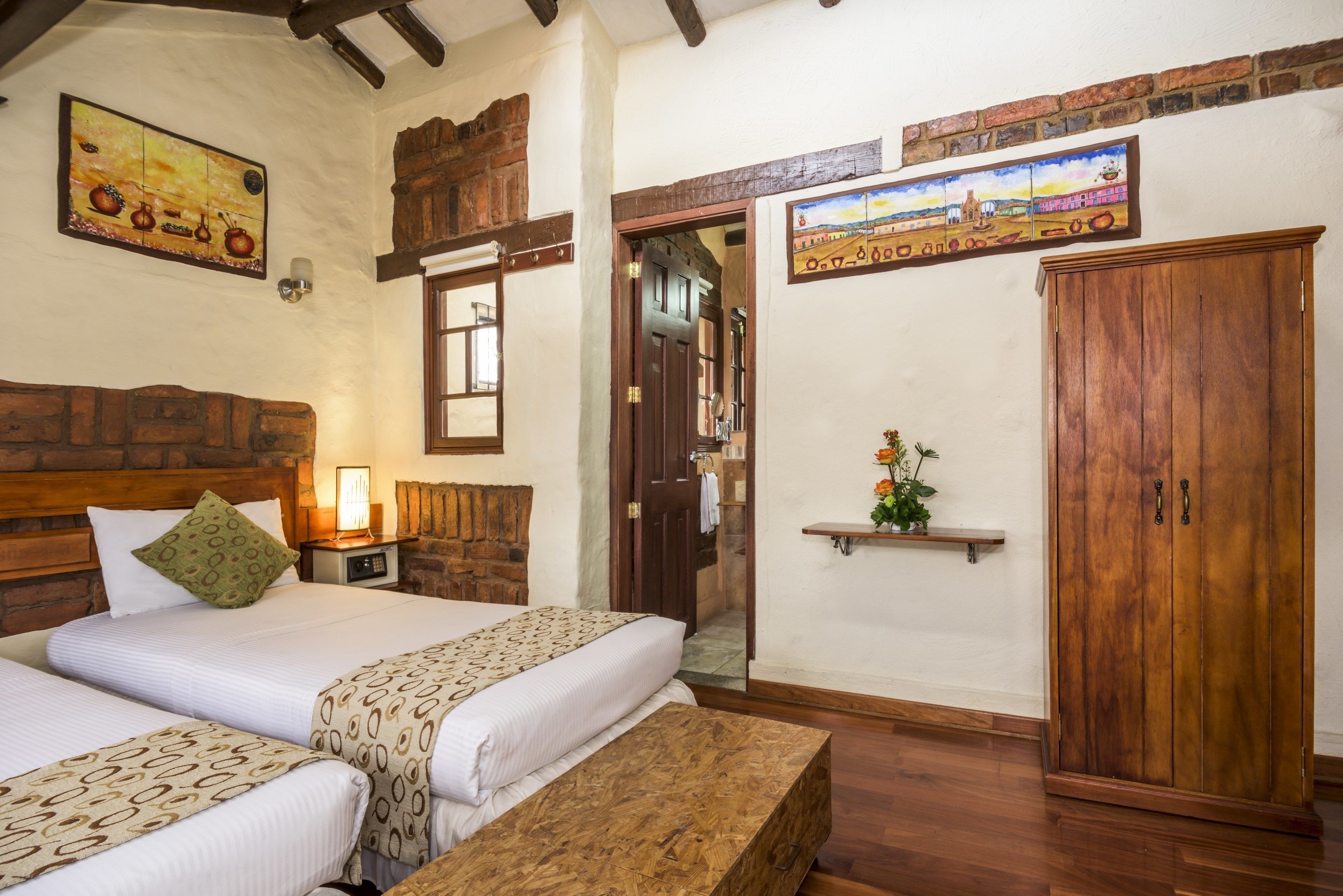 A room with two beds, wooden headboards, a brick wall, a wooden wardrobe, and a small shelf with flowers.