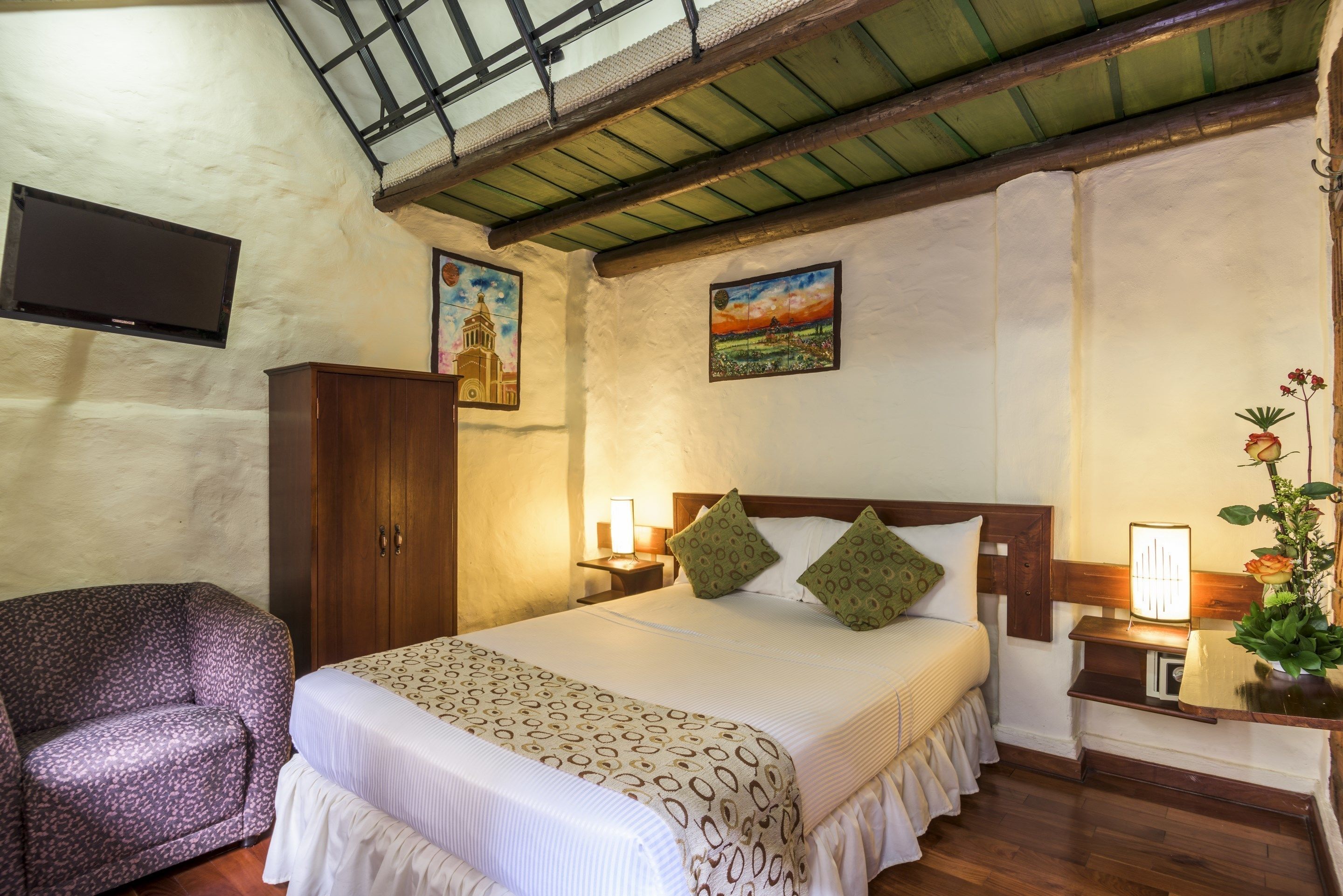 A bedroom with a bed, a wooden wardrobe, a nightstand with a lamp, and a painting on the wall.