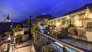 An outdoor terrace with a metal railing, potted plants, and a view of a hill with a church and a building with illuminated patterns. - Hotel Muisca (Bogotá)