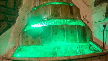 A wooden sauna with a green-lit interior, a carved wooden handle, and a stone wall backdrop.