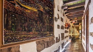 A narrow stone corridor with a large framed painting of a cityscape. The corridor has stone walls, wooden beams, and a tiled floor. - Hotel Muisca (Bogotá)