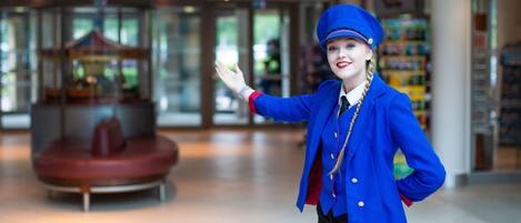 Een persoon in uniform, blauw van kleur, staat in een ruime binnenruimte met een draaimolen en een groot raam op de achtergrond.