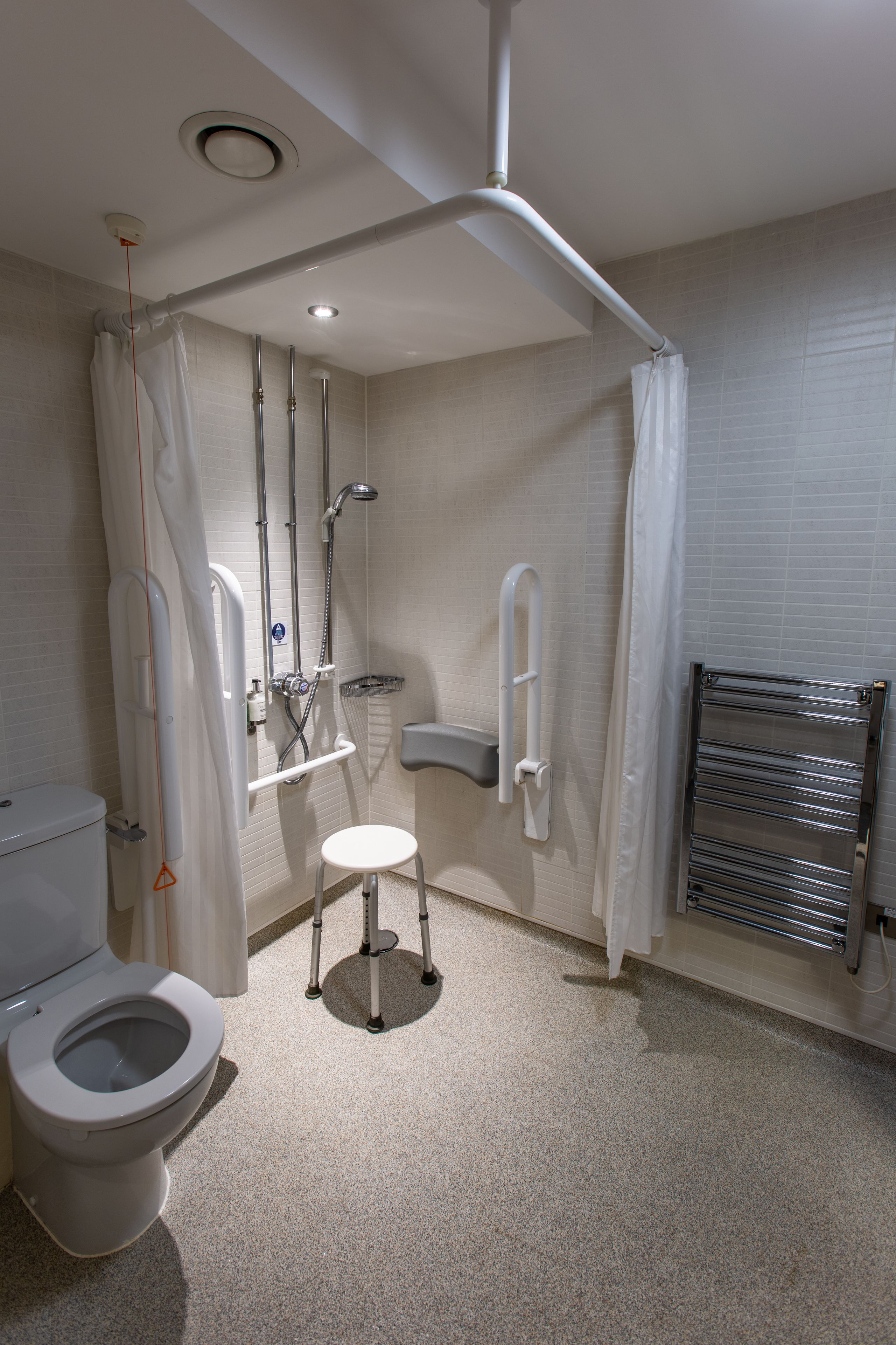 Un baño con inodoro, ducha con barra de apoyo, taburete y toallero.