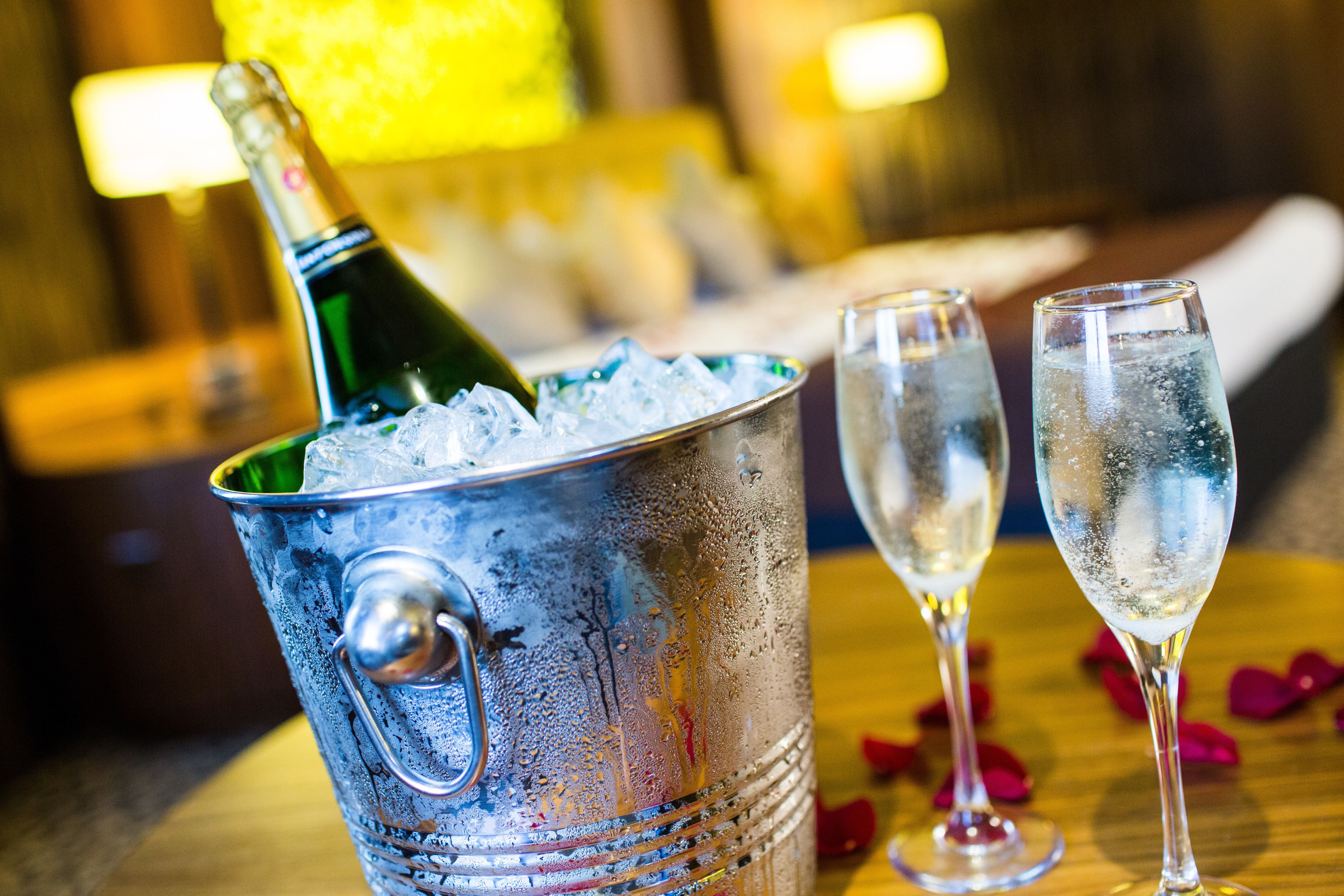 A metallic ice bucket with a bottle of champagne and two glasses filled with champagne, placed on a wooden table with rose petals.