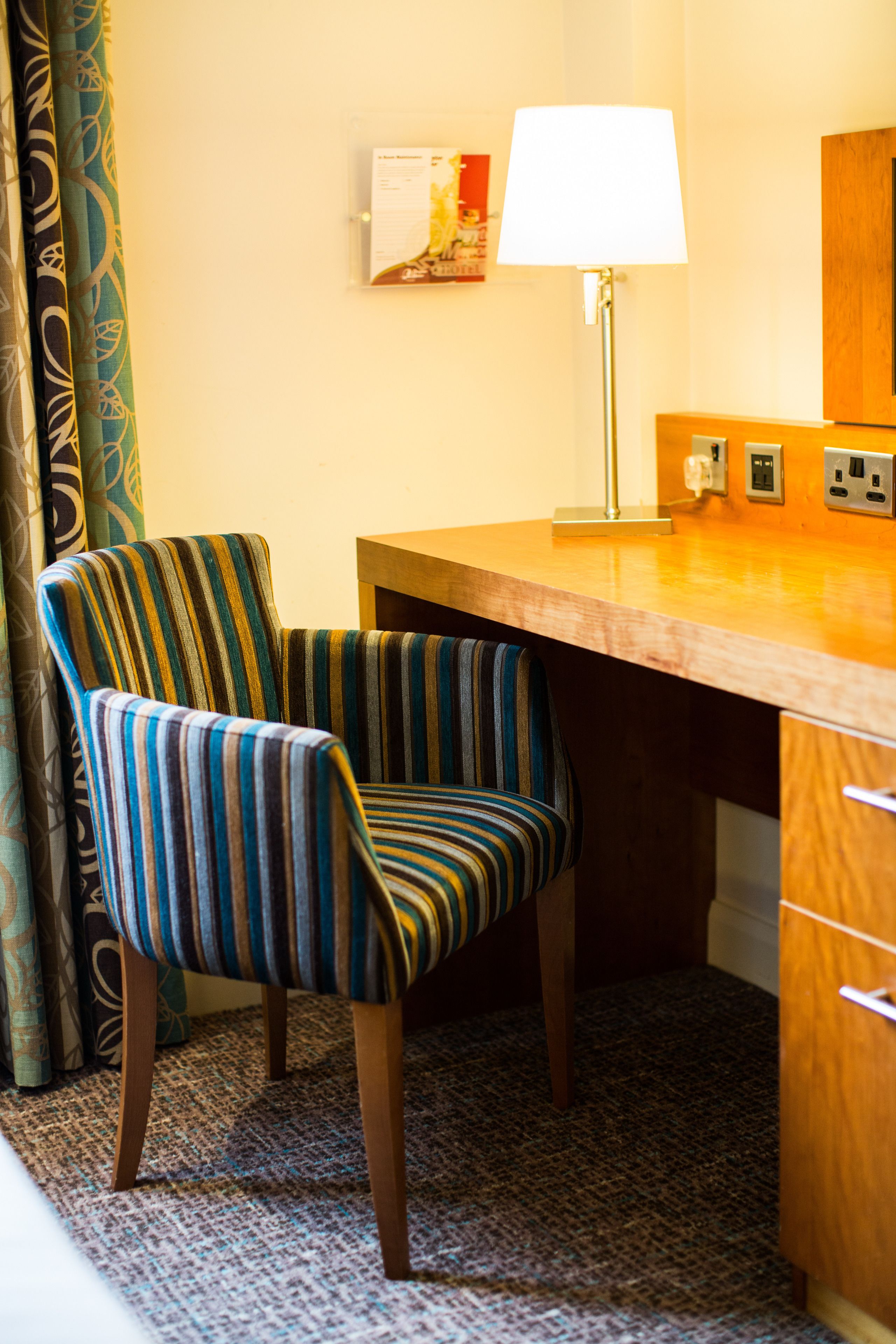 A striped armchair, a wooden desk, a lamp, and a wall-mounted picture in a hotel room.