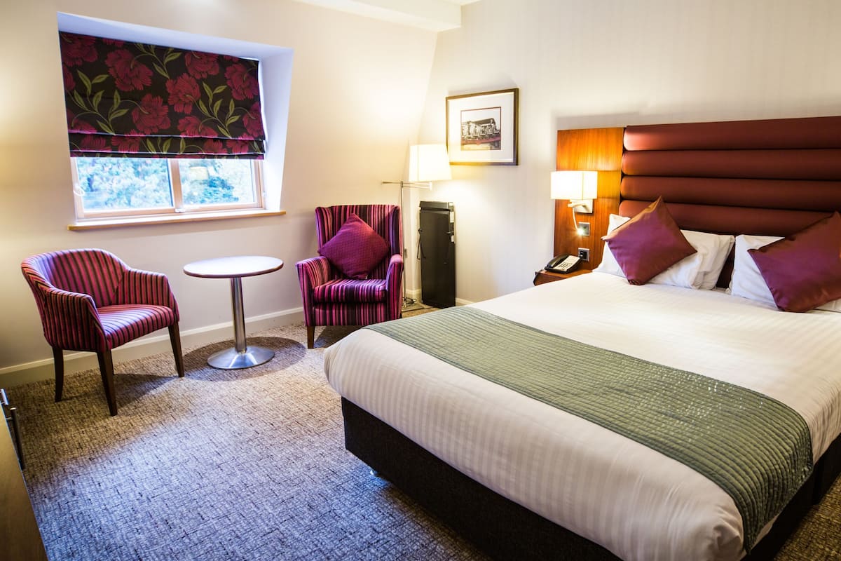 A hotel room with a bed, two armchairs, a small table, a lamp, and a window with a floral blind.