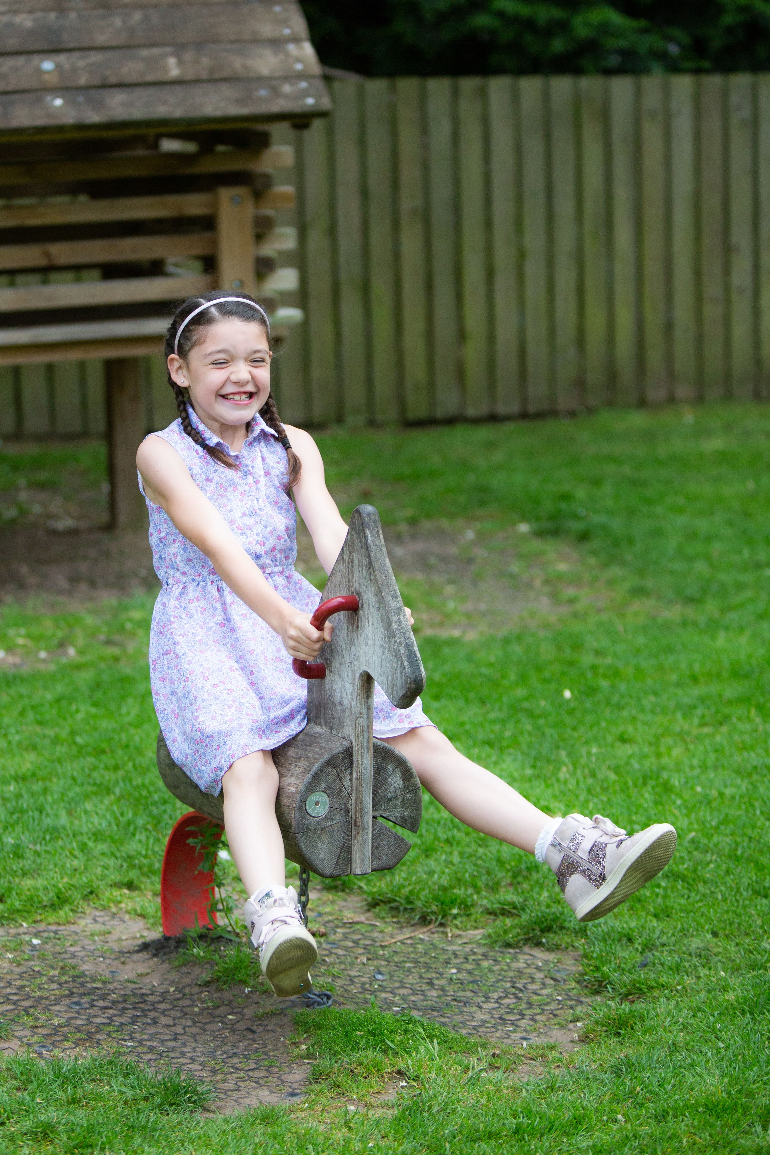 A child on a wooden swing set in a grassy area with a wooden fence in the background.