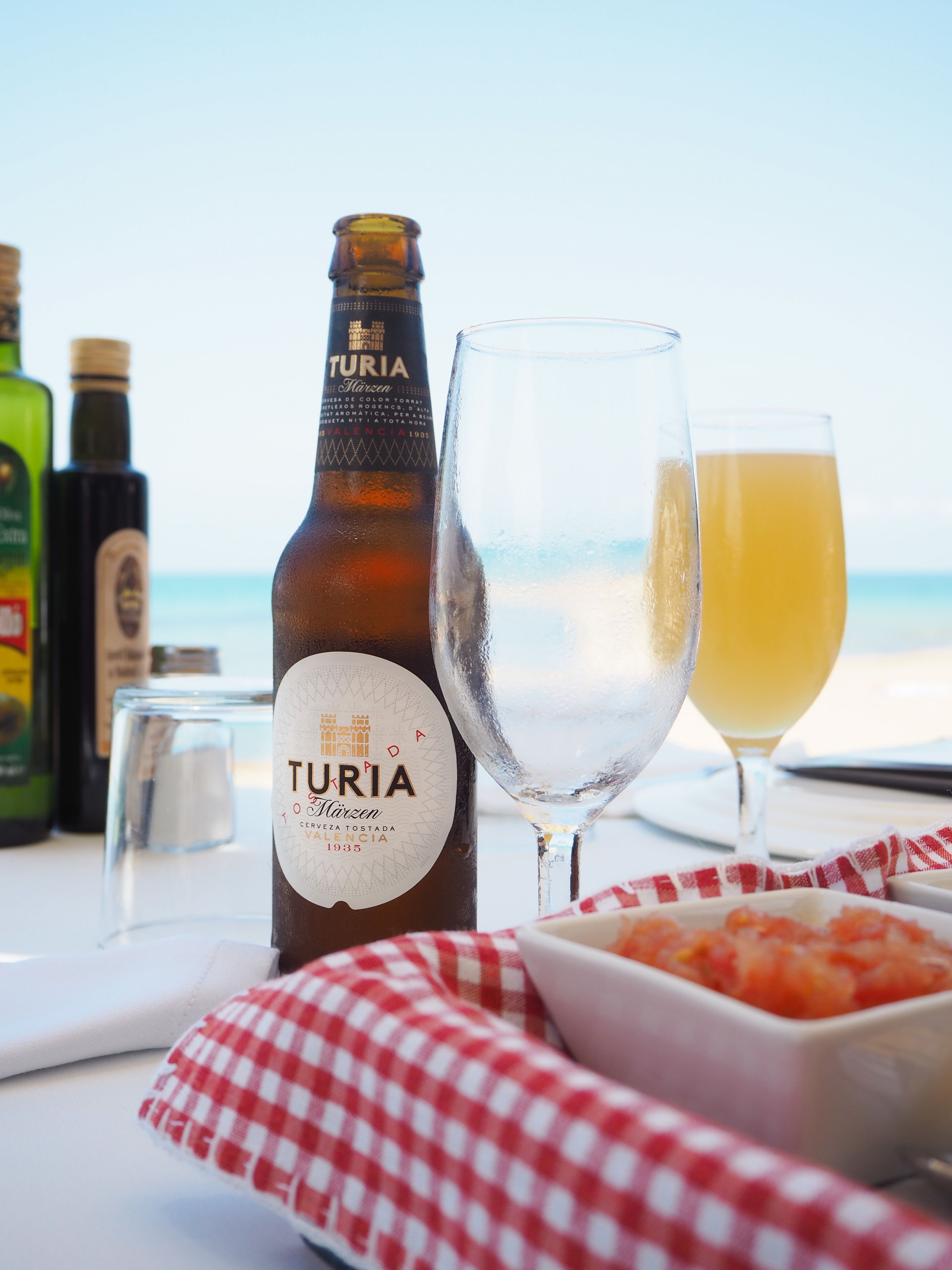 Une bouteille de bière Turia, un verre de bière et un verre de jus d’orange sont posés sur une table, accompagnés d’une serviette à carreaux rouges et blancs et d’un bol de nourriture.