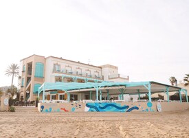 A beachfront hotel with a covered outdoor seating area, multiple balconies, and a mural with aquatic themes.