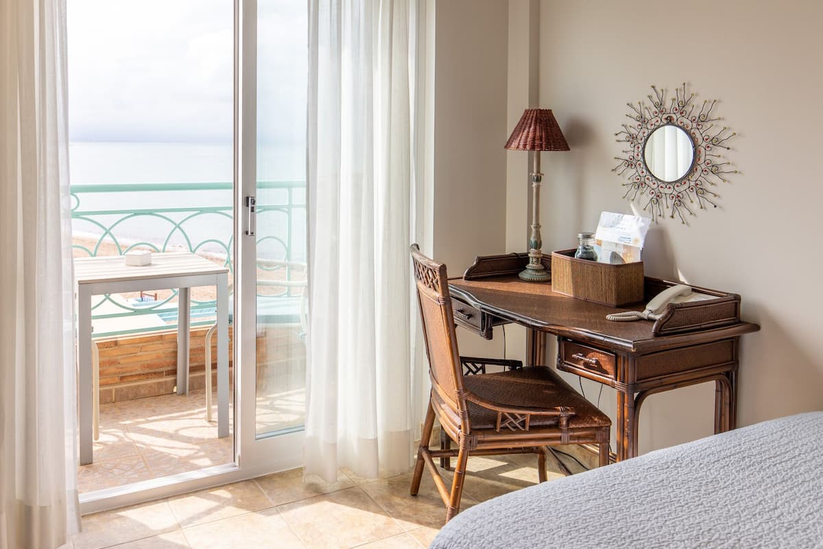 A room with a wooden desk, wicker chair, and a view of the beach through a glass door.