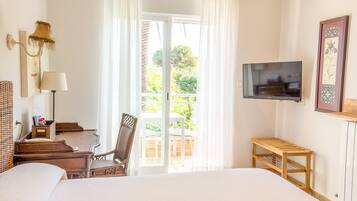 A hotel room with a bed, a desk with a chair, a television, and a balcony with a view of greenery.