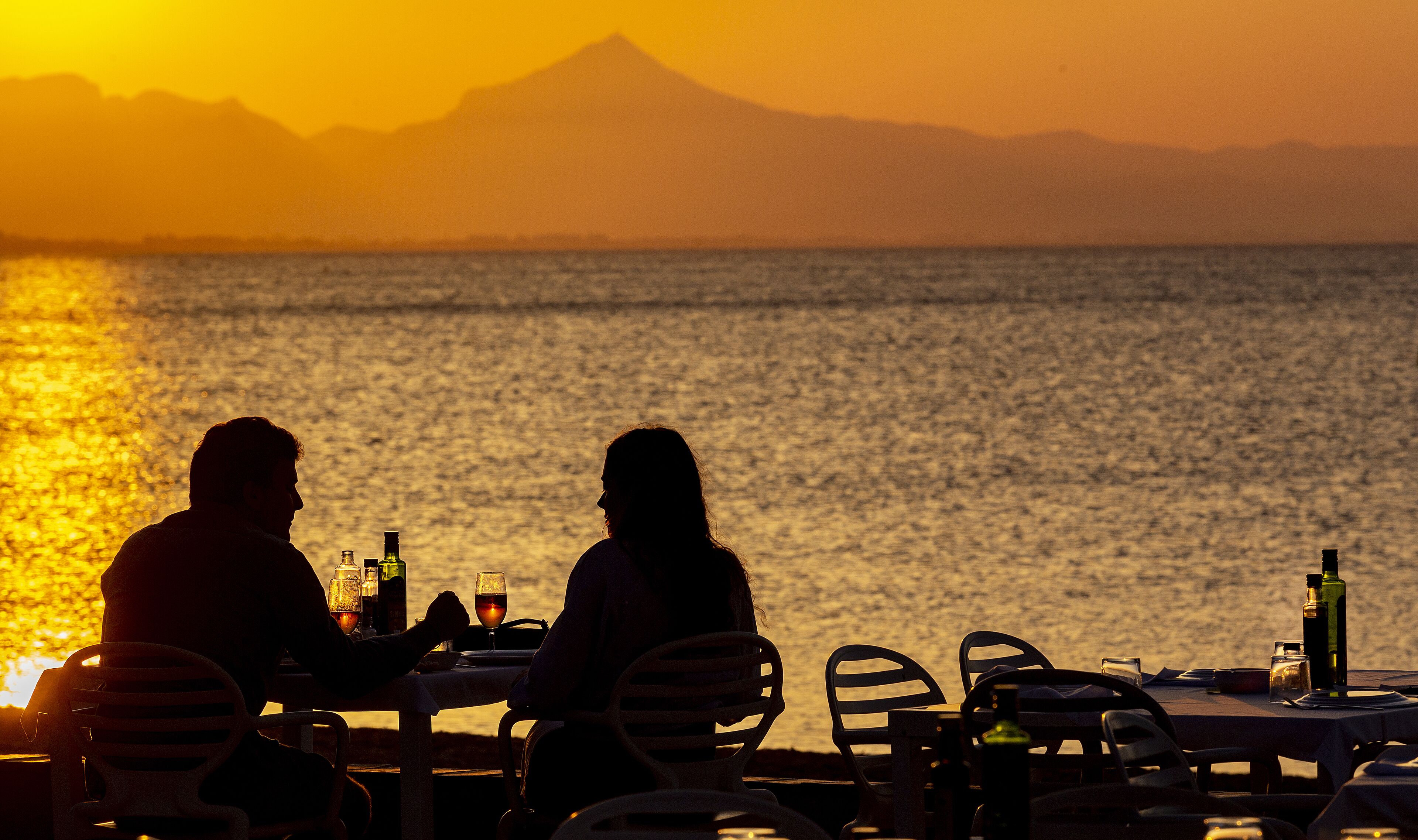 A couple dining at a table with a view of the sea and a mountain in the background.