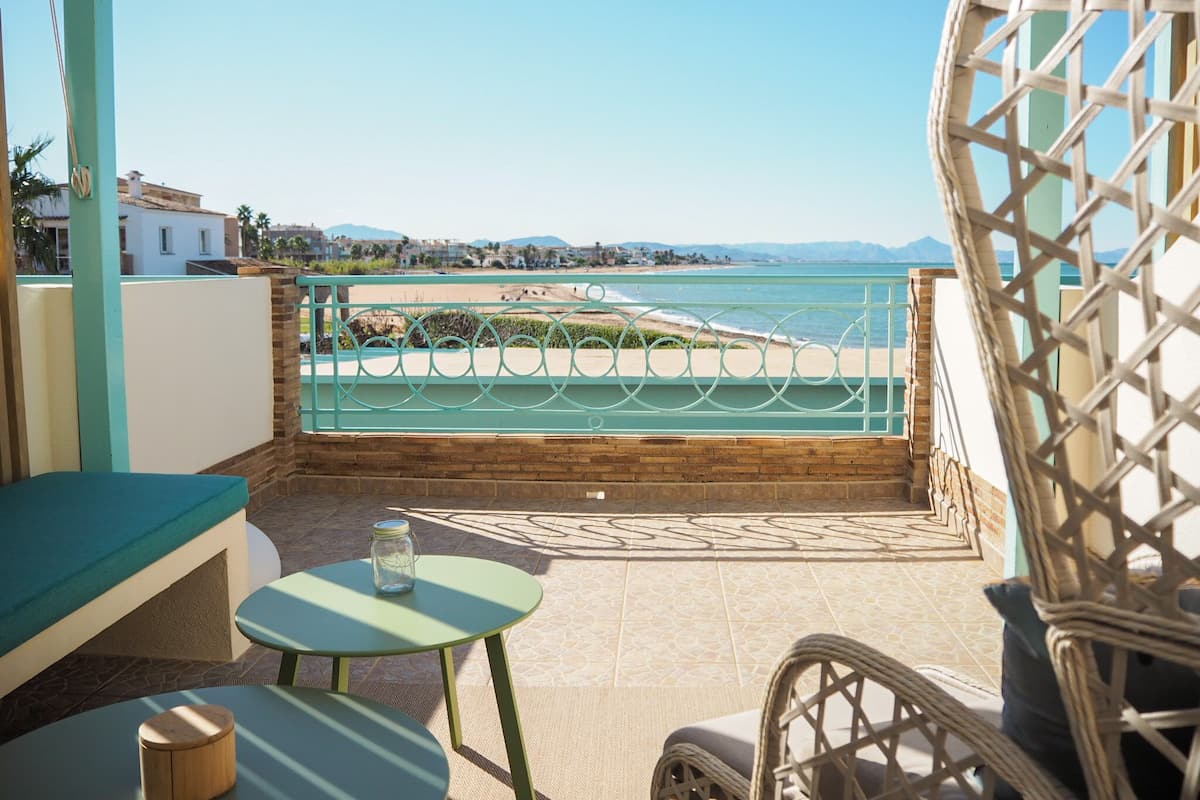 A balcony with a view of the beach and mountains, a wicker chair, a small table, and a cushioned bench.