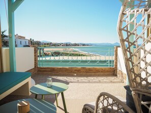A balcony with a view of the beach and mountains, a wicker chair, a small table, and a cushioned bench. - Noguera Mar Hotel (Denia)