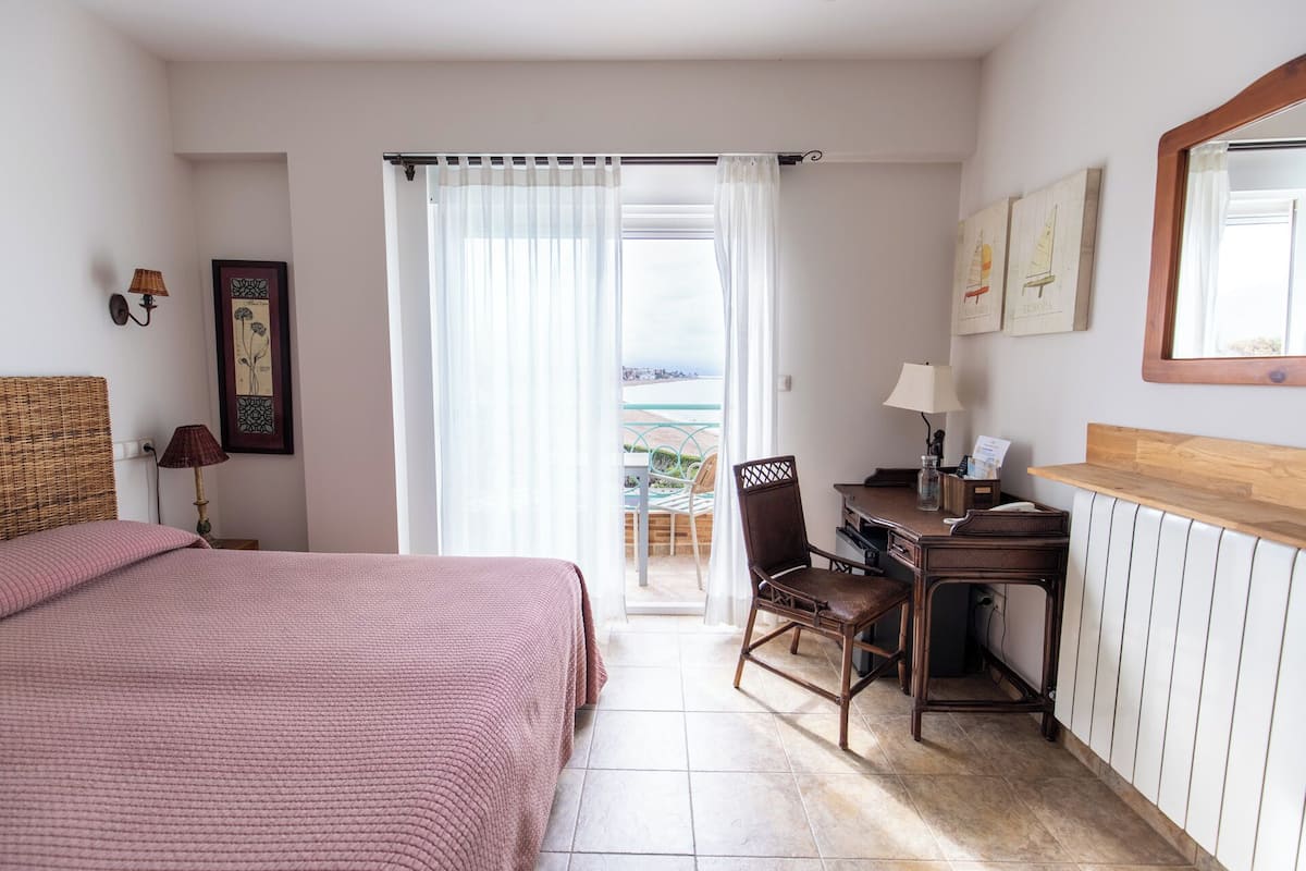 A bedroom with a bed, a desk, a chair, and a view of the sea through a window.