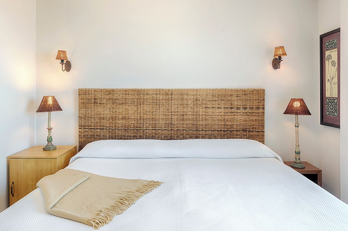 Una cama bien hecha con un cabecero tejido, dos lámparas de noche y una obra de arte enmarcada en la pared.