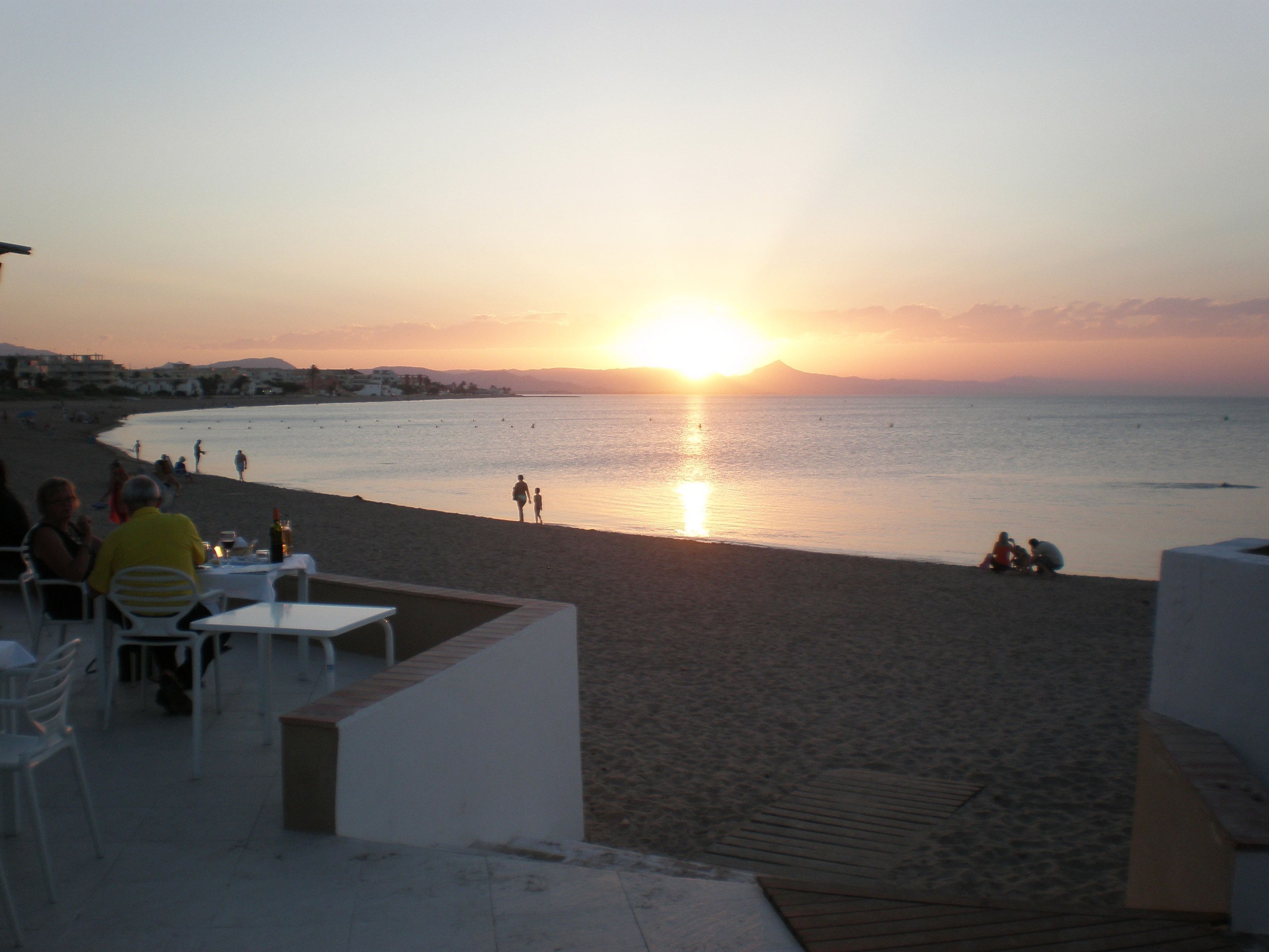 Ein Restaurant am Strand mit Terrassenplätzen und Meerblick, im Hintergrund ein Sonnenuntergang, während Menschen die Aussicht genießen.