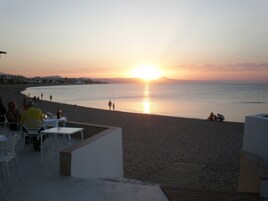 Ein Restaurant am Strand mit Terrassenplätzen und Meerblick, im Hintergrund ein Sonnenuntergang, während Menschen die Aussicht genießen.