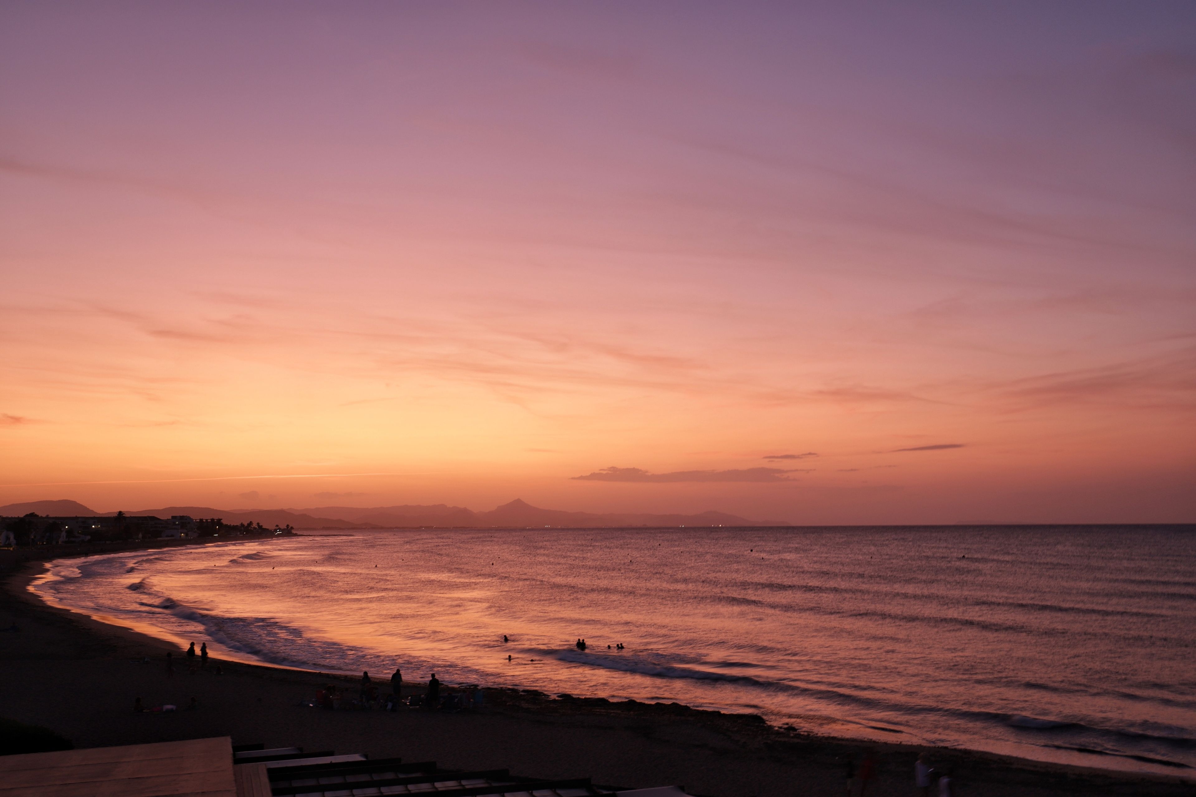 Ein Strand bei Sonnenuntergang mit einer gewundenen Küstenlinie, Personen im Sand und fernen Bergen.