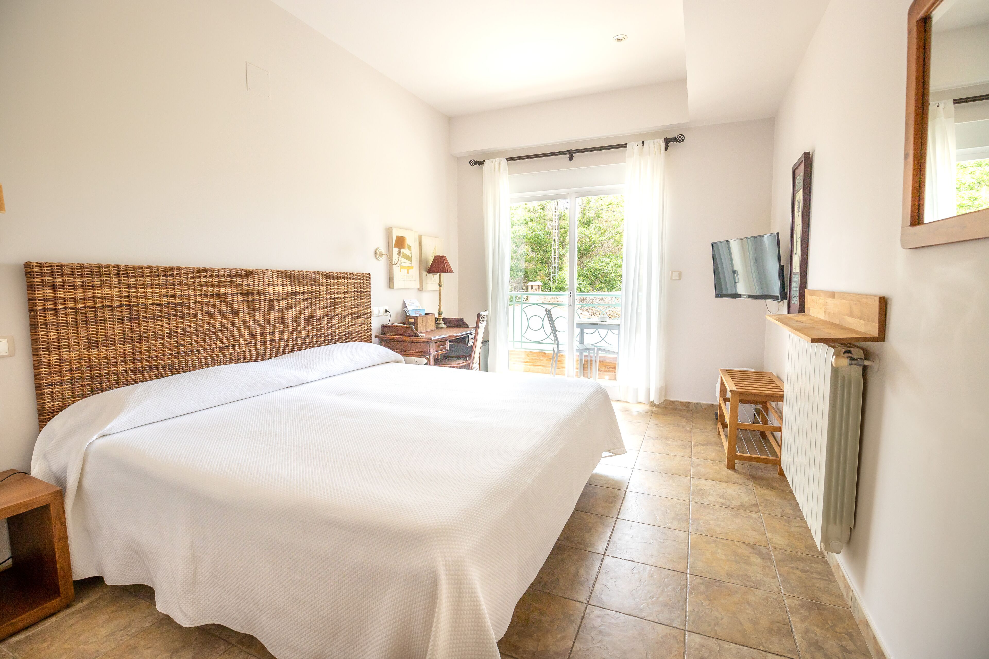 Een slaapkamer met een groot bed, een houten hoofdbord, een klein tafeltje, een stoel, een televisie en een balkon met uitzicht op groen.