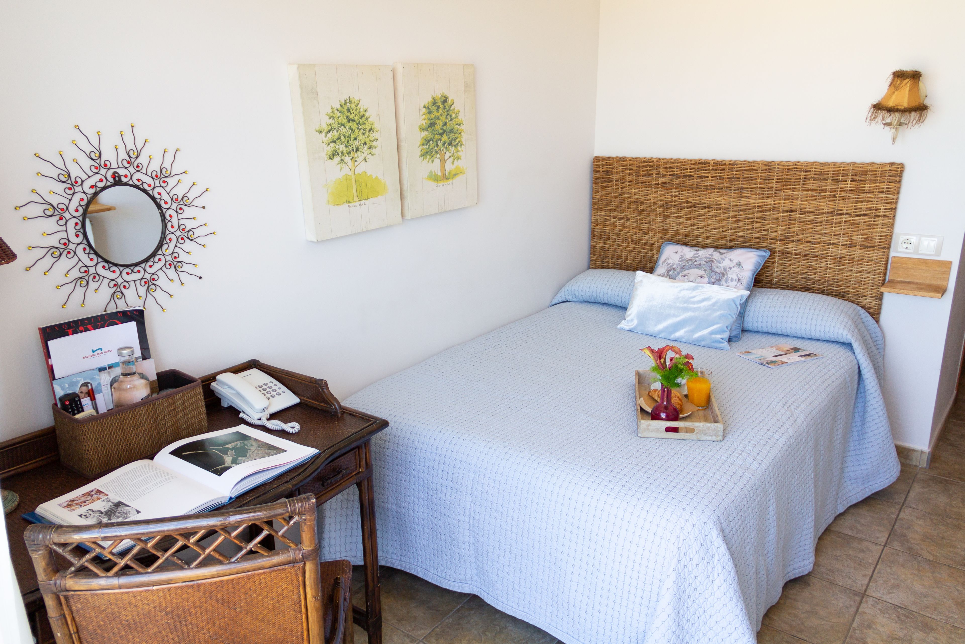 Un dormitorio ordenado con cama, una mesita de noche con un libro y un teléfono, una silla y un espejo montado en la pared.