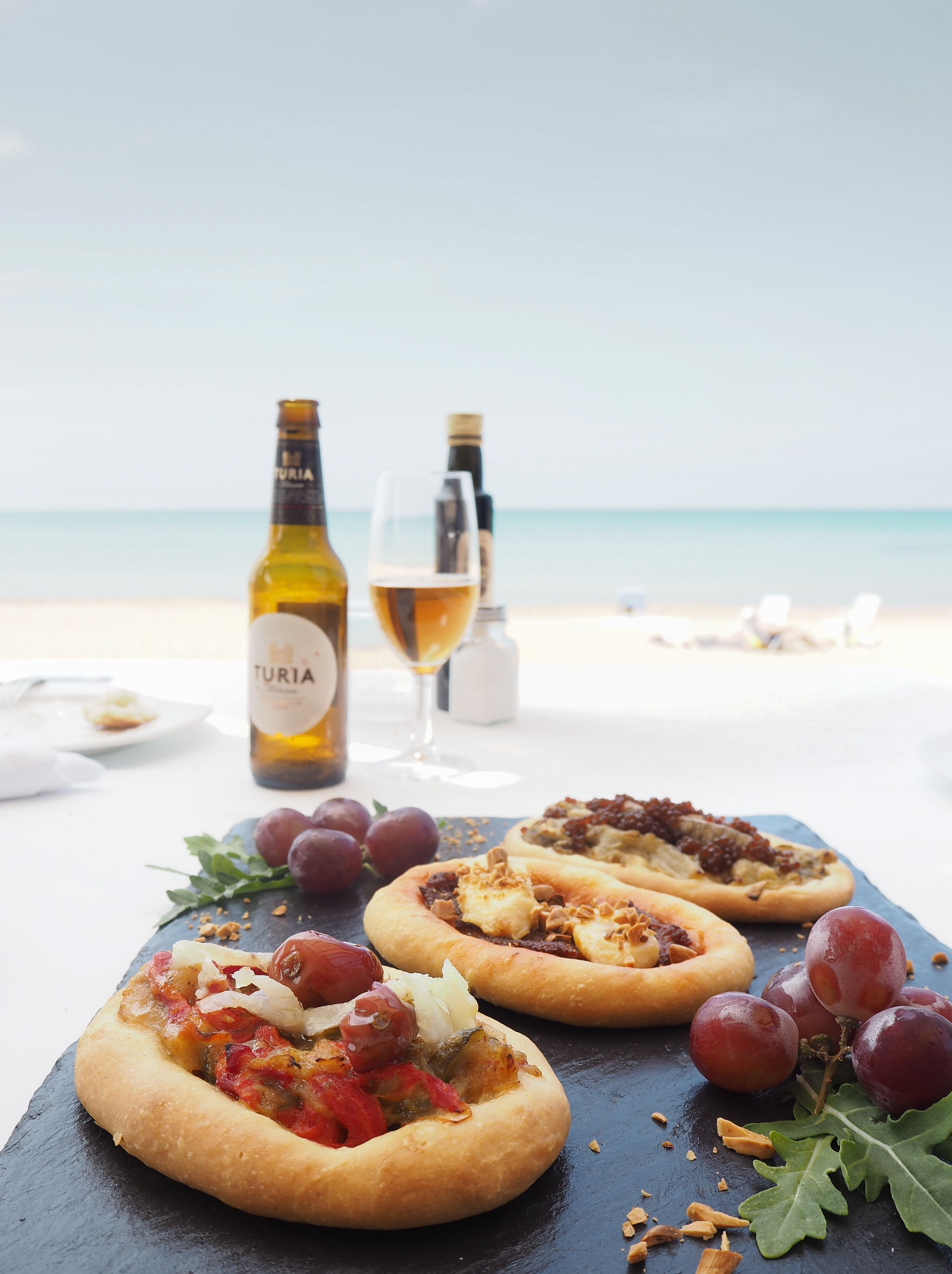 Una taula amb dues pizzes, una ampolla de vi i un got de vi a la platja.