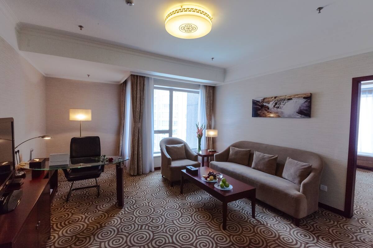 Habitación de hotel con un escritorio de vidrio, dos sillones, un sofá, un televisor de pantalla plana, alfombra estampada, un cuadro en la pared y una ventana con cortinas.