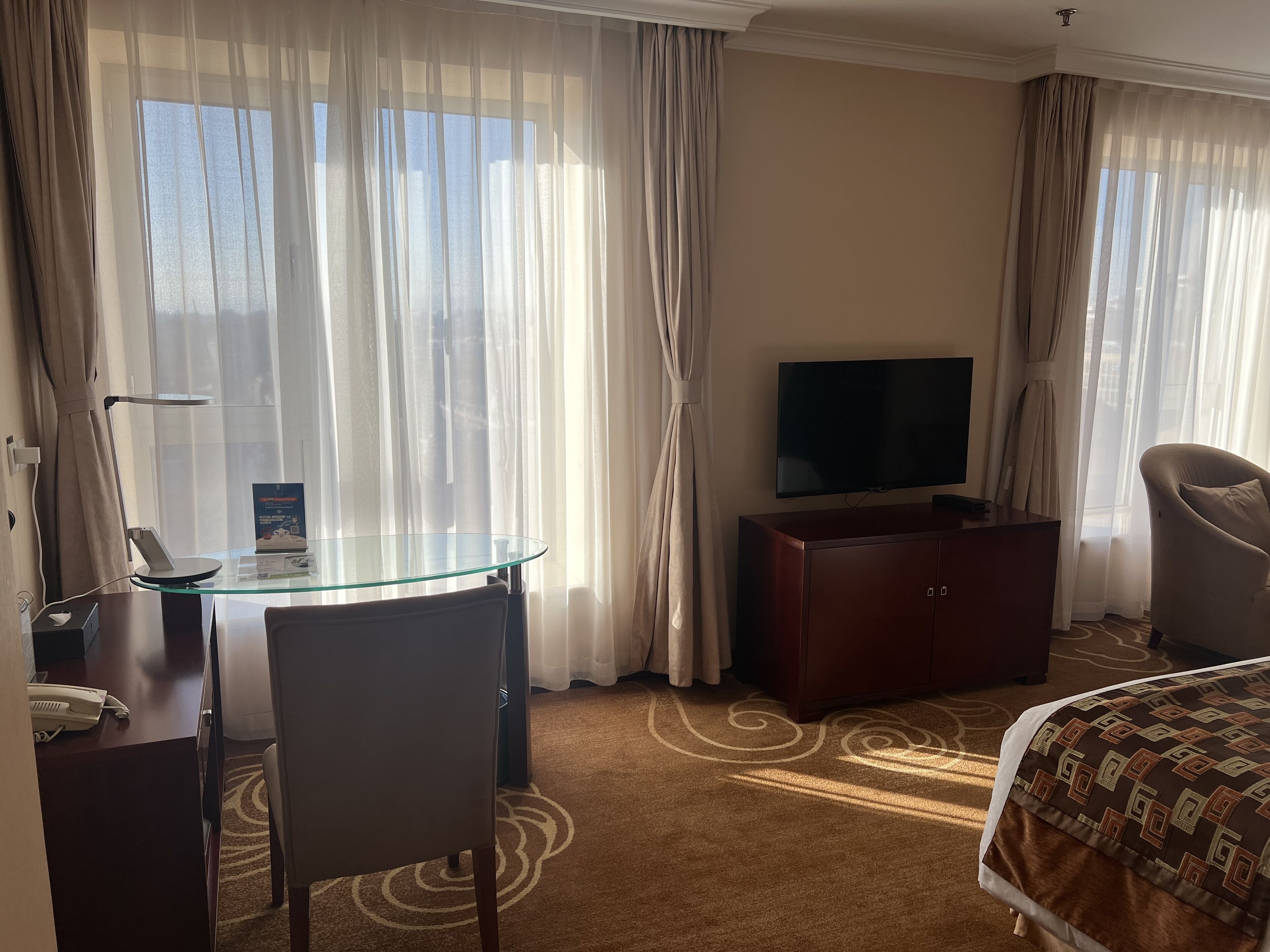 Quarto de hotel com escrivaninha de vidro, televisão sobre um móvel de madeira, cama com roupa de cama estampada e cortinas leves.