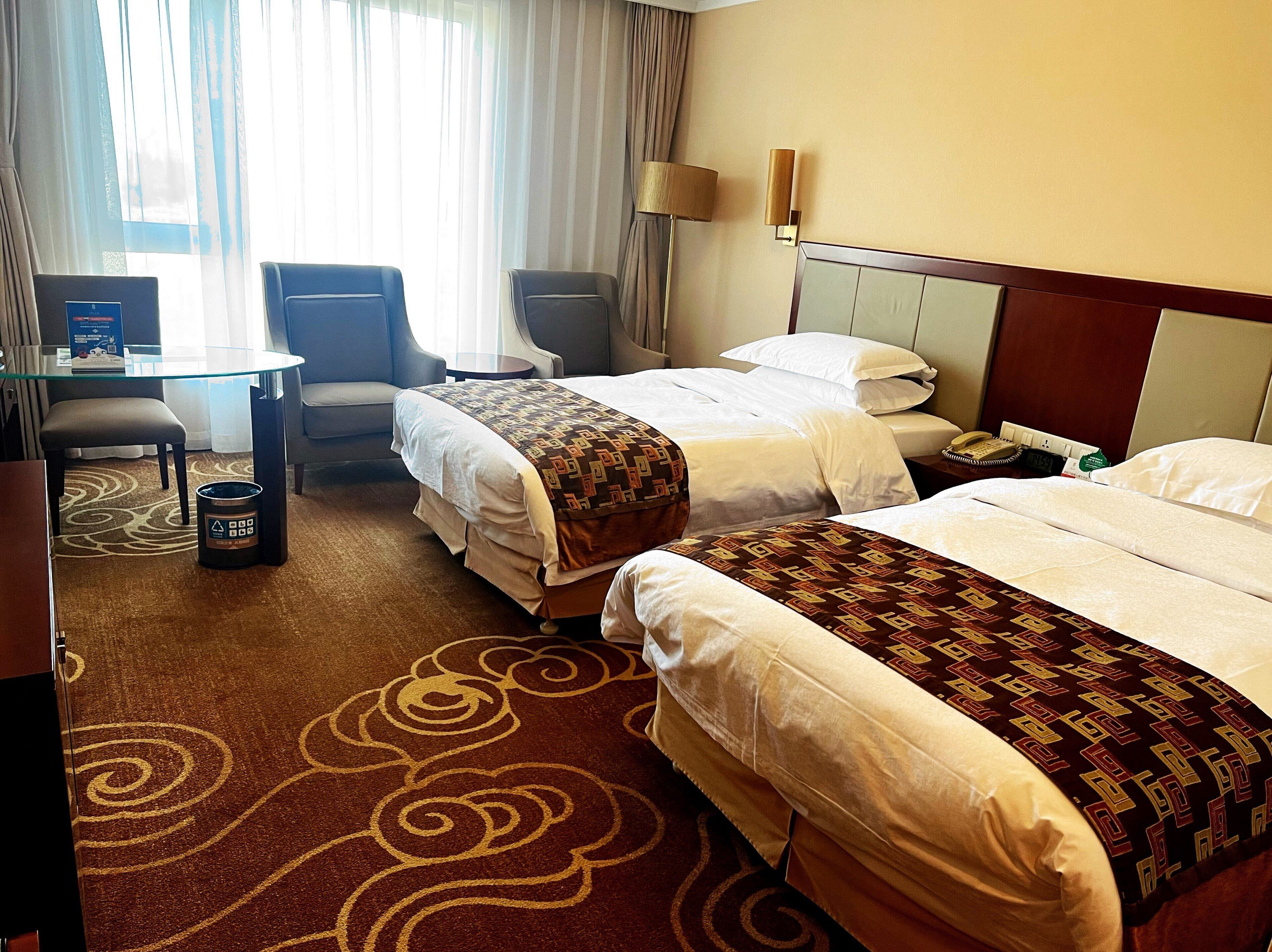 A hotel room with two beds, a desk, a chair, a lamp, and a trash bin.