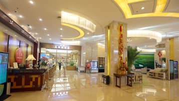 A hotel lobby with a reception desk, a digital screen, a large decorative column, and a seating area with a view of a green landscape.