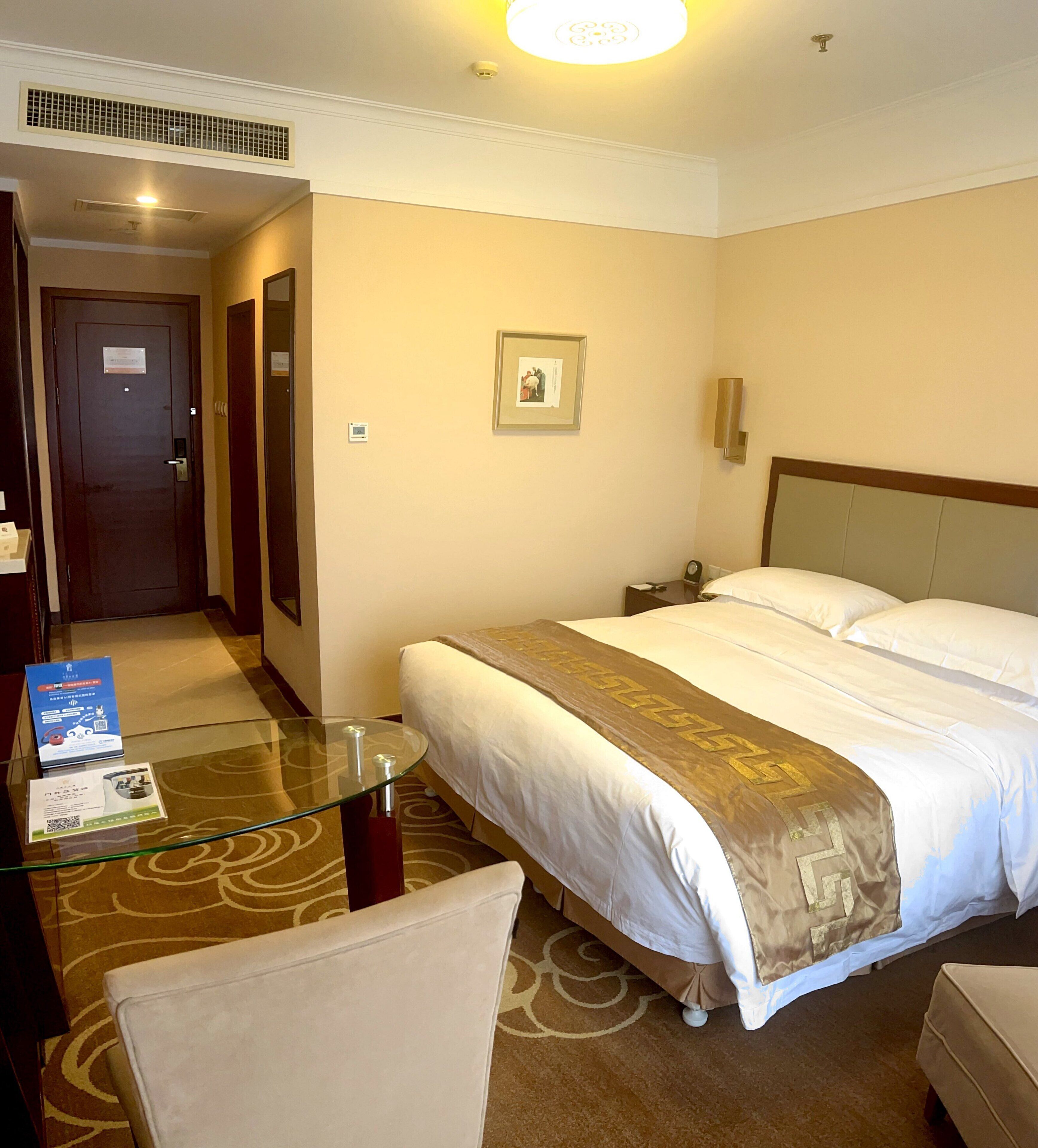 A hotel room with a bed, a glass table, a chair, a nightstand, and a door.