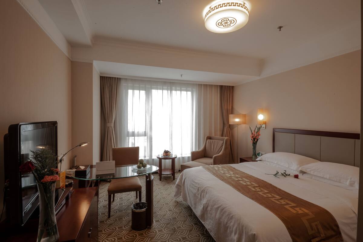Una habitación de hotel con cama, escritorio, silla, televisor y ventana con cortinas.