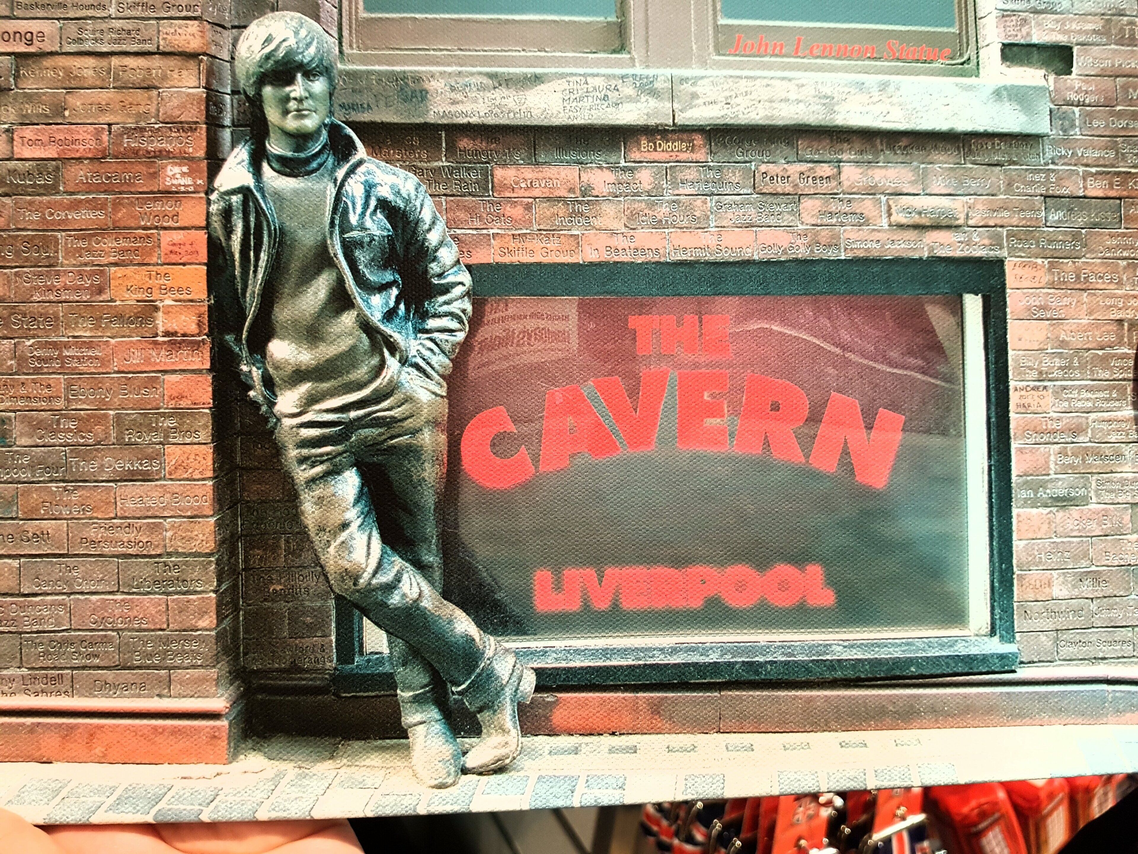 En staty som föreställer en person som lutar sig mot en tegelvägg, och bredvid statyn finns en skylt med texten "The Cavern Liverpool".
