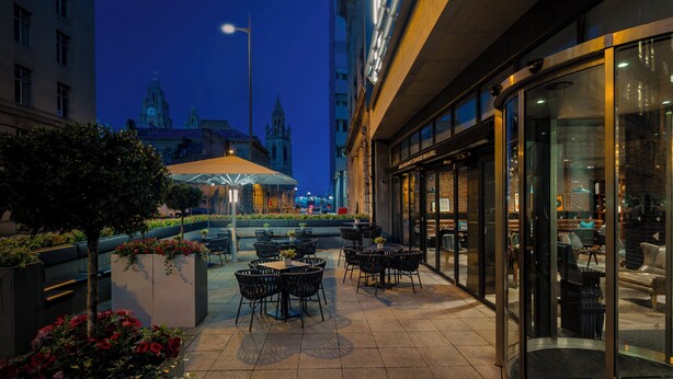 An outdoor seating area with tables and chairs, a planter with flowers, and a view of a historic building with towers in the background.