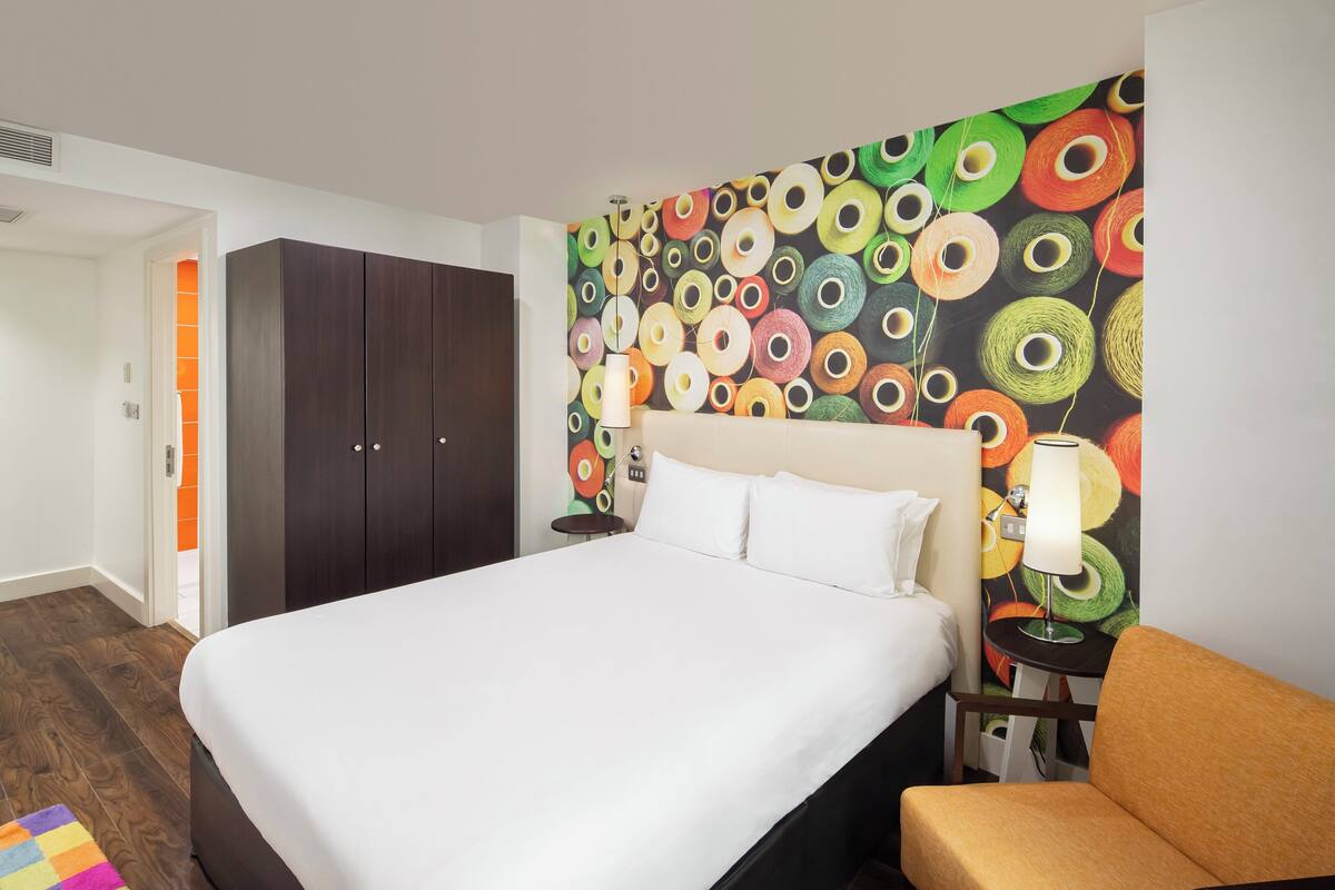 A hotel room with a large bed, a nightstand, a lamp, a wardrobe, and a colorful wall.