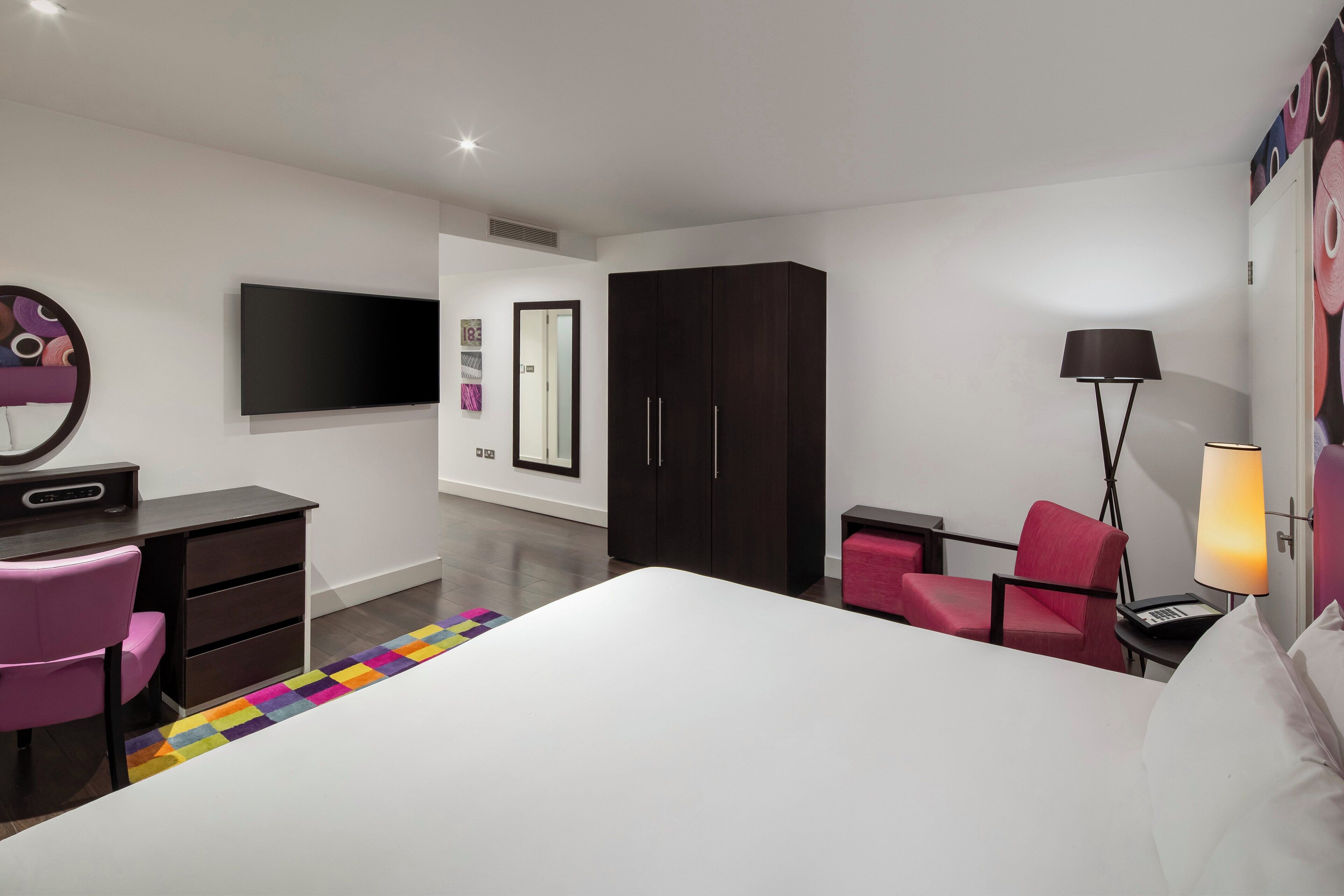Una habitación de hotel moderna con una cama grande, un escritorio con silla, un armario, un televisor y una lámpara.