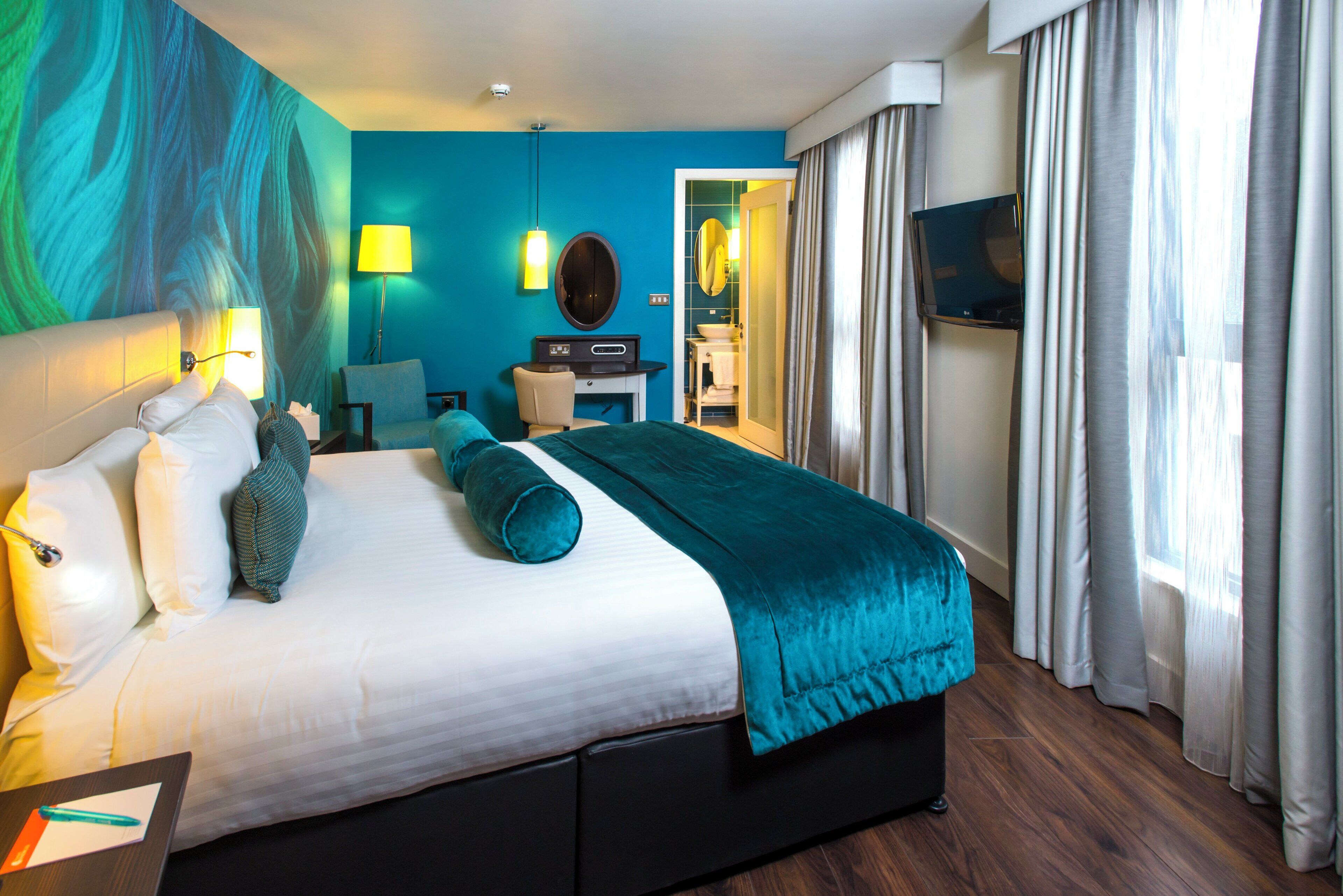 Habitación de hotel con cama, escritorio, silla, televisor y baño visible a través de una puerta abierta.