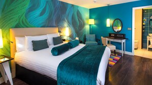 A modern hotel room with a large bed, two bedside tables with lamps, a desk with a chair, a mirror, and a bathroom visible through an open door.