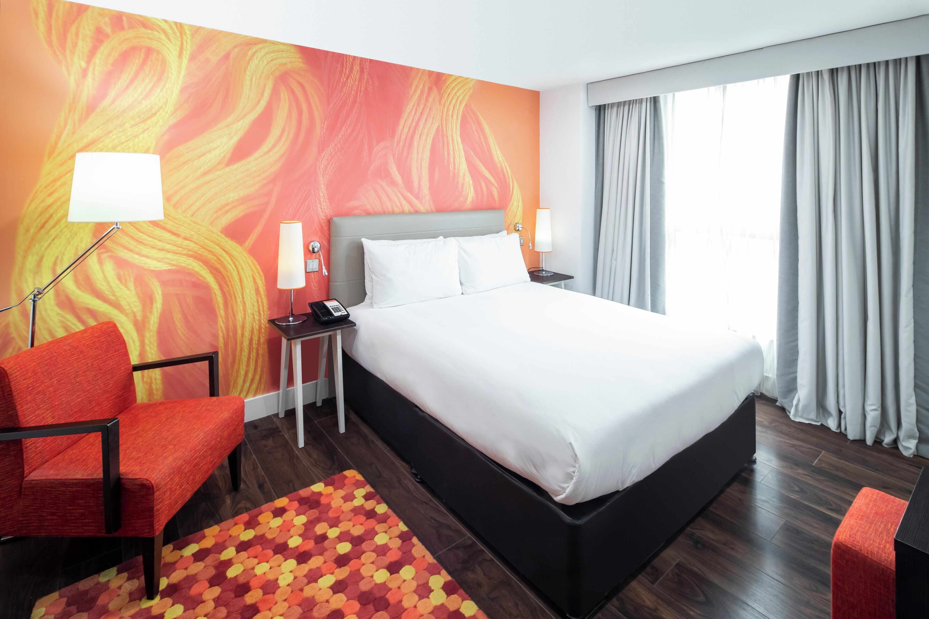A hotel room with a bed, a desk, a chair, a lamp, and a window with curtains.