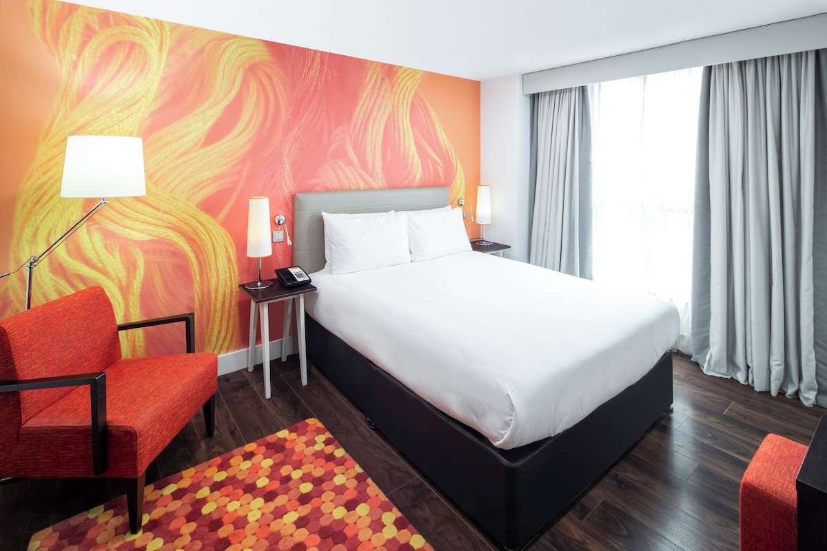 A hotel room with a bed, a desk, a chair, a lamp, and a window with curtains.
