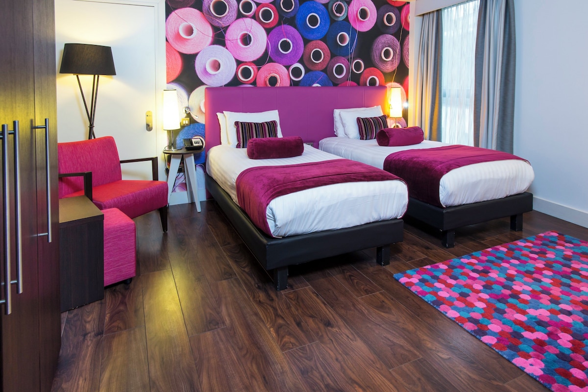 A hotel room with two beds, a desk, a chair, a lamp, and a colorful wall decoration.